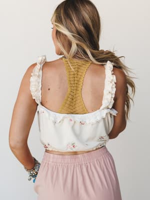 Shot from behind, a model wears a cream floral embroidered crop top with ruffle straps and eyelet trim layered over a mustard lace bralette, paired with a blush pink midi skirt — soft, romantic, and effortlessly put-together.