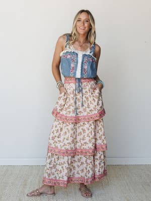 A smiling blonde model wears a floral-embroidered denim crop top with lace trim paired with a tiered floral maxi skirt in cream and pink — the kind of outfit that feels like a summer farmers market and a road trip rolled into one.