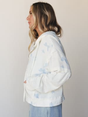 A side-profile shot of a model in a cream zip-up hooded jacket with soft blue butterfly print, styled with wide-leg jeans against a clean studio backdrop — the kind of piece that makes a Saturday morning feel like something worth dressing for.