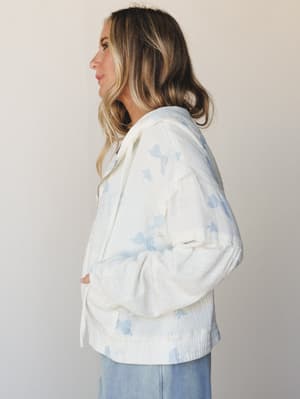 A side-profile shot of a model in a cream zip-up hooded jacket with soft blue butterfly print, styled with wide-leg jeans against a clean studio backdrop — the kind of piece that makes a Saturday morning feel like something worth dressing for.