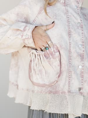 A close-up detail shot of a pink marble-washed denim jacket with a gathered drawstring pocket, crochet hem trim, and snap button closure — her stacked silver and turquoise rings doing just as much styling work as the jacket itself.