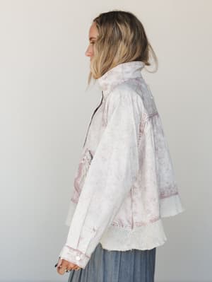 A side-profile shot of a model wearing an oversized acid-wash denim jacket in a soft pink marble wash, layered over what looks like a blue-gray maxi skirt — the crochet hem trim and relaxed silhouette give it that intentionally undone, lived-in feel.
