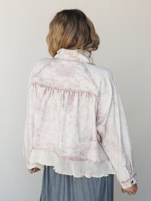 A back-view shot of a relaxed, acid-washed jacket in dusty pink and cream with gathered detailing and open crochet trim at the hem — styled over a soft slate midi skirt for an effortlessly lived-in boho look.