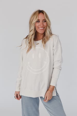 A woman with long blonde hair, wearing the Catch My Smile Pullover Top in khaki and light blue pants, smiles while standing against a plain white background.