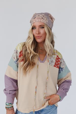 A woman with long, wavy blonde hair wears the Sarah Floral Lace Headscarf in gray, a colorful patchwork sweater, and blue jeans as she stands against a plain light background, looking at the camera with a slight smile.