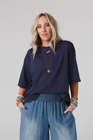 A woman with wavy blonde hair wears The Nest Your Favorite Slub Tee Shirt in navy, wide-leg denim pants, layered necklaces, and colorful beaded bracelets, standing hands-in-pockets against a plain light background.