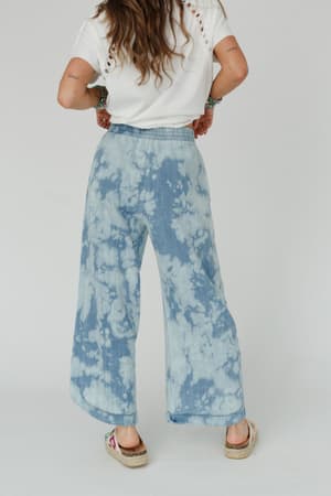 A person stands facing away, wearing The Nest So Comfy Wide Leg Denim Pant in Bleach Dye Denim with a white shirt and casual shoes. Their long hair is down, arms show tattoos and bracelets. Background is plain white.