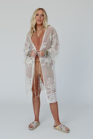 A woman with long blonde hair wears a peach romper, layered necklaces, bracelets, floral slide sandals, and the Butterfly Beach Kimono - Ivory, standing against a plain white background.