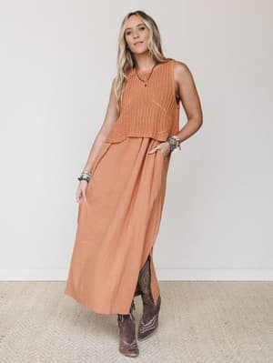A warm, earthy maxi dress in pumpkin orange is layered with a matching sleeveless crochet crop top, styled with brown cowboy boots and stacked turquoise bracelets — the kind of outfit that feels effortless but clearly thought through.