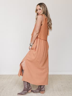 A warm pumpkin-toned maxi dress with a crochet bodice and flowy gauze skirt is styled from the back, paired with dusty mauve cowboy boots and stacked boho bracelets — earthy, effortless, and very much going somewhere fun.