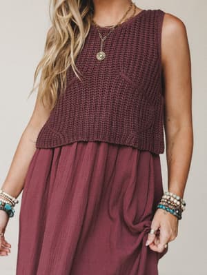 A cropped knit sweater vest layered over a flowy gauze midi dress, both in a deep wine tone — styled with gold layered necklaces and stacked beaded bracelets for that effortless, jewelry-everywhere kind of boho.