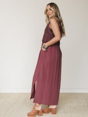 A warm, earthy look shot in a clean studio — she's wearing a wine-toned maxi dress with a crochet overlay top, paired with tan suede clogs and a stack of beaded bracelets that feel like they've been collected over years, not styled in five minutes.