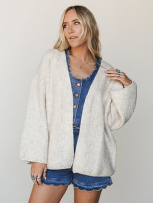 An oversized cream cardigan layered over a denim button-front romper with scalloped edges — the kind of effortless outfit that looks like it came together in two minutes but clearly didn't.