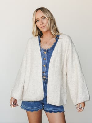 A cream knit cardigan with billowy sleeves is layered over a denim button-front top and scallop-hem denim shorts — the kind of effortless mix that looks thrown together but absolutely isn't.