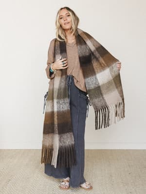 A relaxed full-body shot of a model holding out an oversized plaid fringe scarf in warm earthy tones, layered over a slouchy v-neck tee and wide-leg dark denim — the kind of effortless fall outfit that looks like it came together in two minutes but s