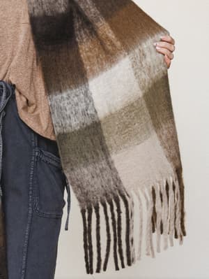 A close-up lifestyle shot of a oversized plaid scarf held out by a model wearing a camel tee and dark cargo pants — the scarf's earthy patchwork of brown, sage, and cream with long fringe trim feels like something you'd grab off a hook on the way out