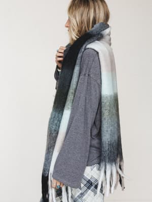 A side-profile shot of a model wrapped in an oversized plaid blanket scarf with long fringe, layered over a heathered gray long-sleeve tee and plaid wide-leg pants — the whole look feels like the coziest Saturday morning that somehow still looks inte