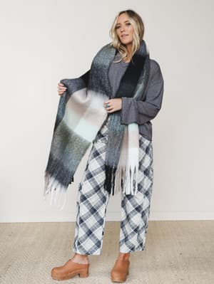 A relaxed, layered fall look built around an oversized plaid blanket scarf — she's holding it open like she's about to wrap herself in it, and honestly same.