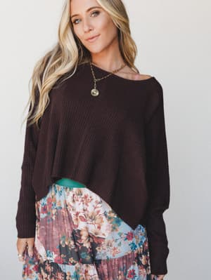 A relaxed ribbed sweater in deep plum-brown is styled over a tiered floral midi skirt, layered with a gold pendant necklace — the kind of effortless mix that looks like it came together in five minutes and still turns heads.