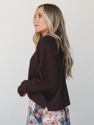 A side-profile shot of a model in a deep plum-brown ribbed cropped sweater paired with a floral skirt peeking at the hem — the kind of cozy-meets-pretty combo that feels effortless on a crisp fall afternoon.