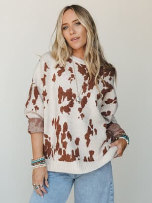 A cozy cow-print knit sweater in cream and camel brown, styled with light wash jeans and layered boho jewelry — the kind of piece that makes a Tuesday feel intentional.