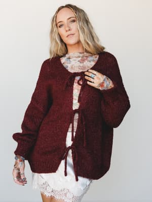 A chunky knit burgundy cardigan tied at the chest and hem is layered over a floral top and white lace mini skirt — the kind of cozy, collected outfit that feels like it came together effortlessly but absolutely didn't.