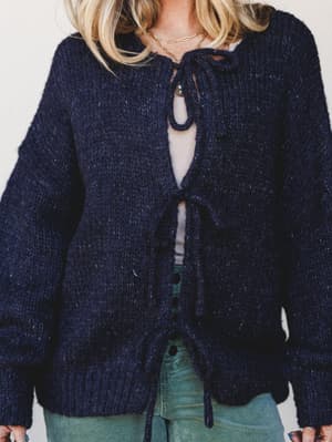 A chunky navy knit cardigan with tie-front closures layered over a sheer top and sage green corduroy pants — the kind of sweater that makes you want to sit near a window with something warm to drink.