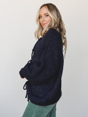 A chunky navy knit sweater with tie details photographed from the side on a model glancing back over her shoulder — the kind of sweater that makes you want to cancel plans and stay in, but make it look intentional.