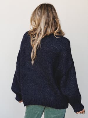 Shot from behind, a model in an oversized navy knit sweater paired with sage green corduroy pants — the kind of cozy-cool combo that makes staying in feel like a choice.