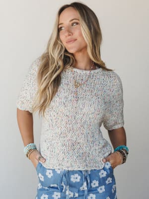 A cropped short-sleeve knit sweater in oatmeal with multicolor confetti speckles, styled with blue floral print pants and layered beaded jewelry — the kind of outfit that feels like a good mood in textile form.