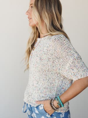 A side-profile shot of a model in a relaxed open-knit sweater top speckled with confetti-like multicolor flecks, styled with floral blue shorts and stacked beaded bracelets — the kind of easy, happy outfit that feels like a good day already.