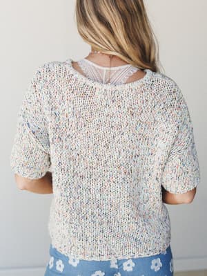 Shot from behind, a model wears a chunky open-knit sweater in creamy oatmeal flecked with confetti-like multicolor speckles — layered over a lace cami and paired with a periwinkle floral skirt, giving the whole look a soft, playful springtime feel.