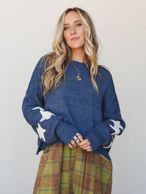 A relaxed knit sweater in heather navy with cream star appliqués on the sleeves, styled over a plaid midi skirt and layered with silver and turquoise rings and a coin pendant necklace — the kind of outfit that feels like autumn in the best possible w