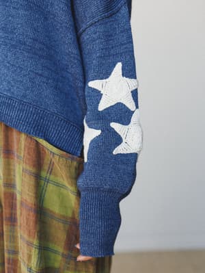 A close-up detail shot of a heather navy knit sweater sleeve with chunky crochet star appliqués, layered over a warm plaid midi skirt — the kind of outfit that feels like a mood board come to life.