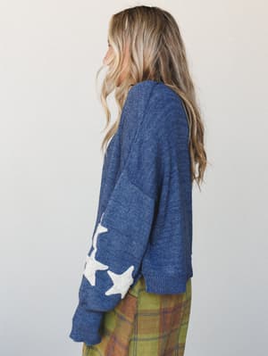 A side profile shot of a star-print knit sweater in heather navy, styled with a plaid skirt — cozy, a little whimsical, and exactly the kind of thing you reach for when the weather turns.