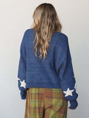 Back view of a relaxed knit sweater in heathered navy with chunky white star appliqués along the sleeves, paired with olive and rust plaid wide-leg pants — cozy and a little dreamy, like something you'd wear to a bonfire and never want to take off.