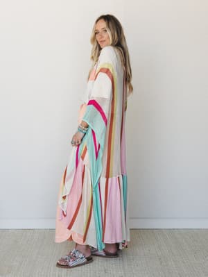 A long open-front knit kimono in cream with bold multicolor stripe rays draped over a peach maxi dress — the kind of layering piece that turns a simple outfit into something worth remembering.