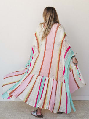 A floor-length open-weave kimono in a vivid multicolor stripe swirls mid-movement, shot from behind — all the joy of a beach coverup and the drama of a maxi duster in one.