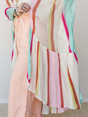 A close-up detail shot of a multicolor striped mesh knit kimono layered over peach wide-leg pants, styled with stacked bracelets and rings for a free-spirited, colorful look.