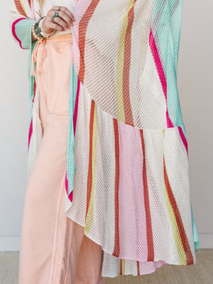 A close-up detail shot of a multicolor striped mesh knit kimono layered over peach wide-leg pants, styled with stacked bracelets and rings for a free-spirited, colorful look.