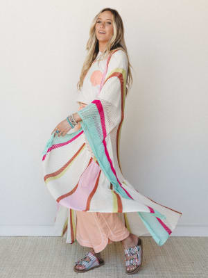 A long-haired model twirls a flowing cream open-knit kimono layered over a graphic tee and peach wide-leg pants — the striped color bursts of hot pink, mint, rust, and chartreuse catching movement like confetti in slow motion.