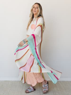 A long-haired model twirls a flowing cream open-knit kimono layered over a graphic tee and peach wide-leg pants — the striped color bursts of hot pink, mint, rust, and chartreuse catching movement like confetti in slow motion.