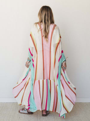 A floor-length open-knit kimono in cream with bold multicolor stripes fans out dramatically from the back, layered over a blush pink dress and paired with stacked bracelets and sandals — the kind of piece that makes a regular afternoon feel like an o