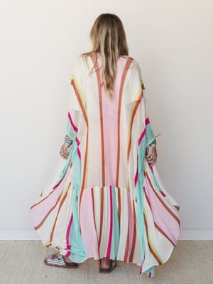 A floor-length open-knit kimono in cream with bold multicolor stripes fans out dramatically from the back, layered over a blush pink dress and paired with stacked bracelets and sandals — the kind of piece that makes a regular afternoon feel like an o