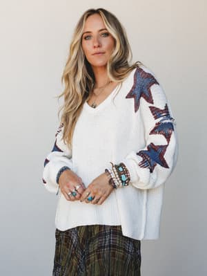 An oversized cream knit sweater with bold star-print sleeves worn over a plaid skirt, styled with layers of turquoise and silver jewelry — the kind of outfit that feels intentional without trying too hard.