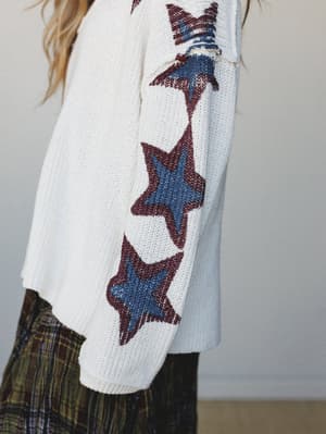 A close-up of an oversized cream knit sweater with bold star intarsia running down the sleeve — layered over a plaid skirt for a vintage-leaning, effortlessly expressive look.