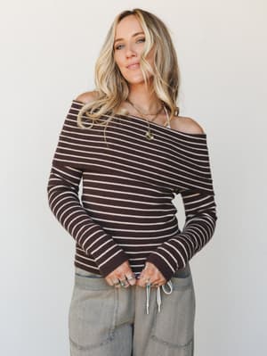 A ribbed, off-shoulder long-sleeve top in deep chocolate with thin cream stripes, styled with olive wide-leg pants and layered gold jewelry — the kind of effortless combo that looks like you tried just enough.