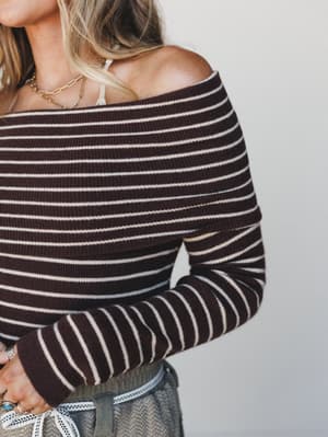 A close-up of a ribbed off-shoulder striped sweater top in deep chocolate and cream, styled with layered gold necklaces and a peek of olive bottoms — the kind of top that does all the heavy lifting on a casual day.