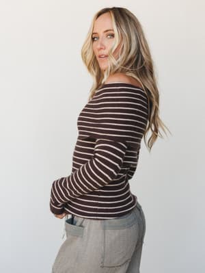 A ribbed striped long-sleeve top in deep chocolate and cream worn off one shoulder, paired with textured gray pants — the kind of effortless combo that looks like you tried just enough.
