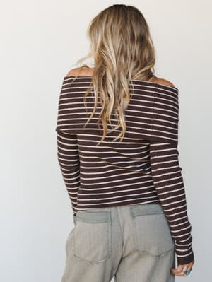 A back-facing shot of a ribbed, off-shoulder striped knit top in deep chocolate and cream, paired with relaxed sage-toned wide-leg trousers and stacked rings — effortlessly collected and just the right kind of cozy.